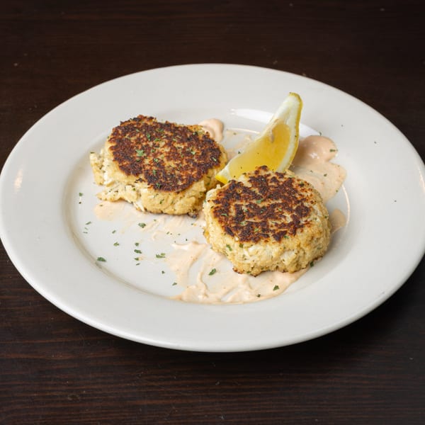 Two golden-brown crab cakes with a lemon wedge and creamy sauce on a white plate.
