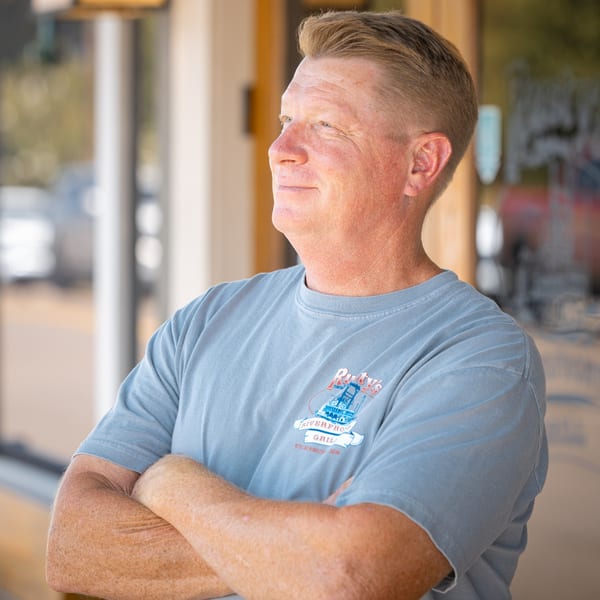 Man in a blue Rusty's Riverfront Grill t-shirt, arms crossed, looking away from the camera.